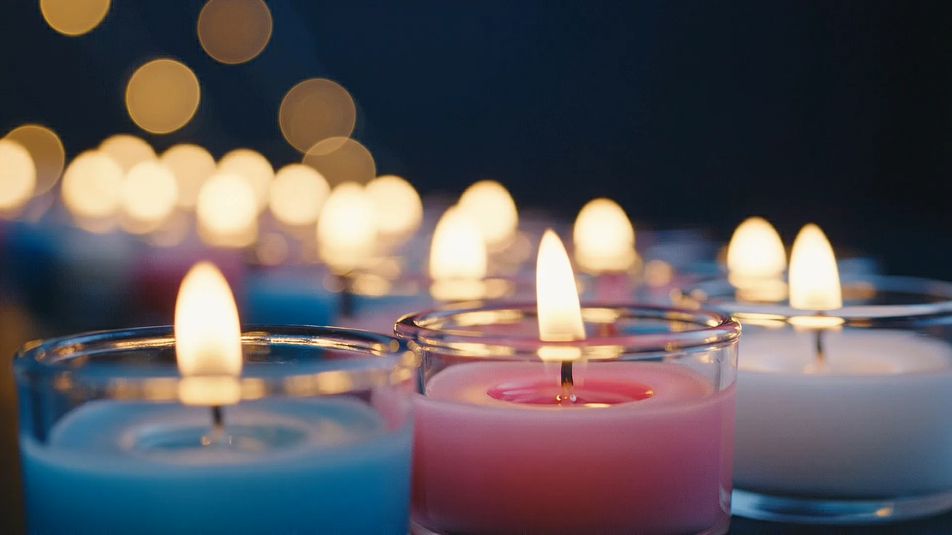 Lit tea light candles in blue, pink, and white colors with bokeh lights in dark background for Transgender Day of Remembrance